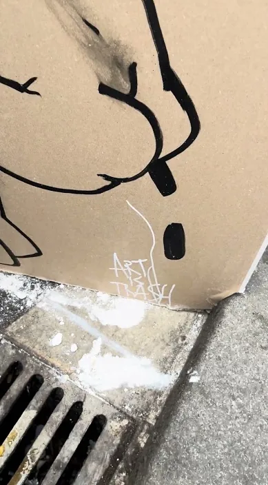 Cartón con un dibujo apoyado sobre una rejilla de desagüe en la calle, Barcelona – Art Is Trash